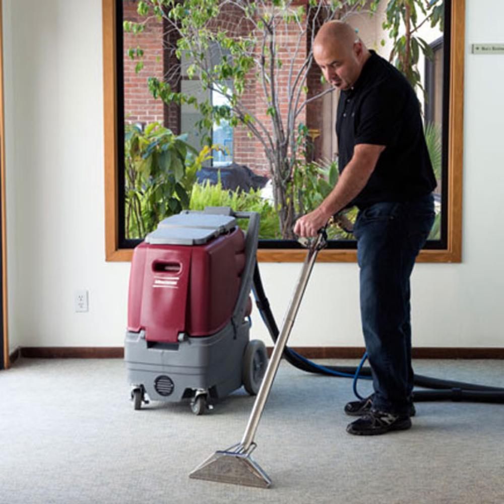 man pushing stainless steel wand attached with a black hose to a red and grey pull along carpet extractor. 