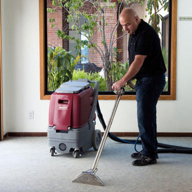 man pushing stainless steel wand attached with a black hose to a red and grey pull along carpet extractor. 