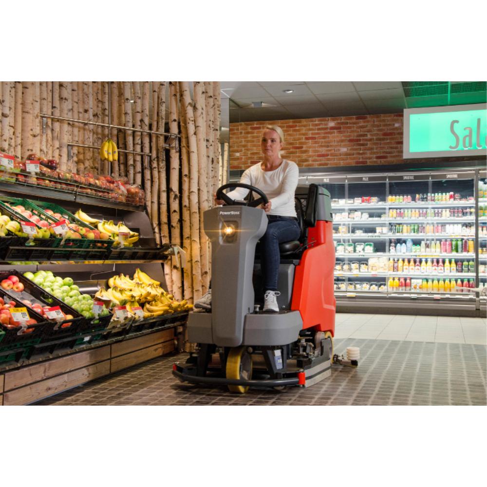 Woman riding a grey and orange floor scrubber in produce of isle of a grocery store.