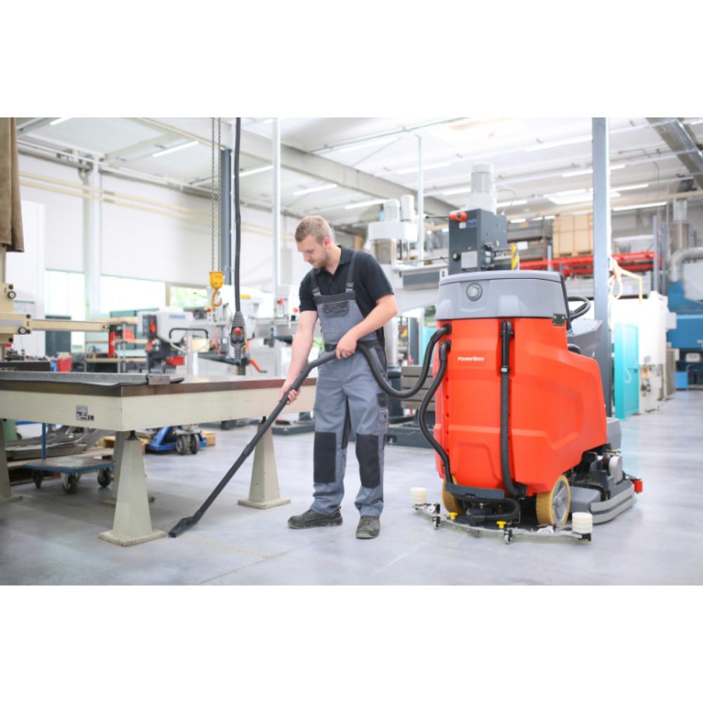 man using vacuum attachment that's connected to grey and orange floor scrubber in a warehouse. 